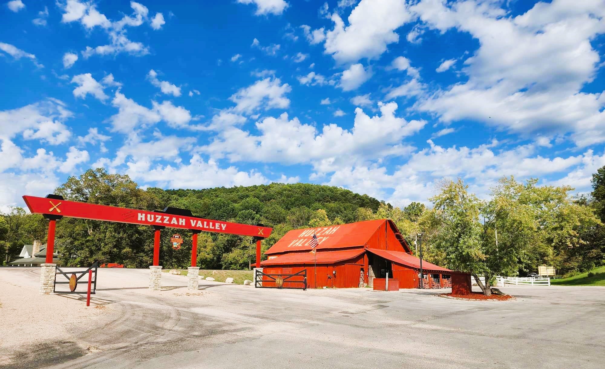A vibrant, sunny landscape featuring the entrance to Huzzah Valley. The scene includes a large red archway with "Huzzah Valley" in bold white letters and a rustic red building to the right. Lush green trees and a cloudy blue sky backdrop complete the setting.