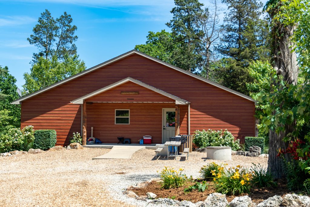 A red-brown wooden cabin with a covered front porch stands amidst lush greenery on a sunny day. The ground is covered with pebbles, and there are various items including chairs, coolers, and tables in front and on the porch. Flowering plants and trees surround the area.
